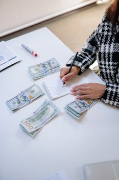 A woman calculating finances with piles of cash and a notepad on a white table.