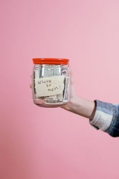 Close-up of a hand holding a savings jar filled with money against a pink backdrop.